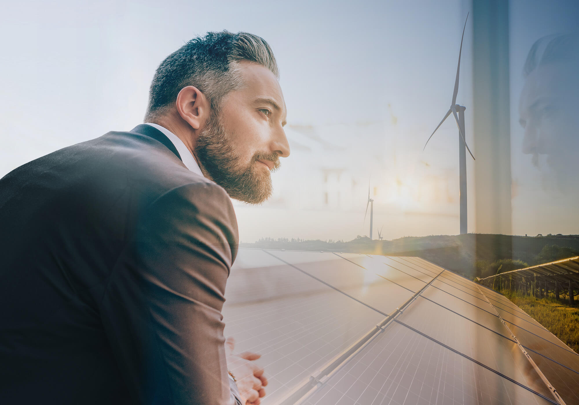 Un homme d'affaires en costume, pensif, qui regarde par une fenêtre. En arrière-plan, des images superposées de panneaux solaires et d'éoliennes au coucher du soleil. 